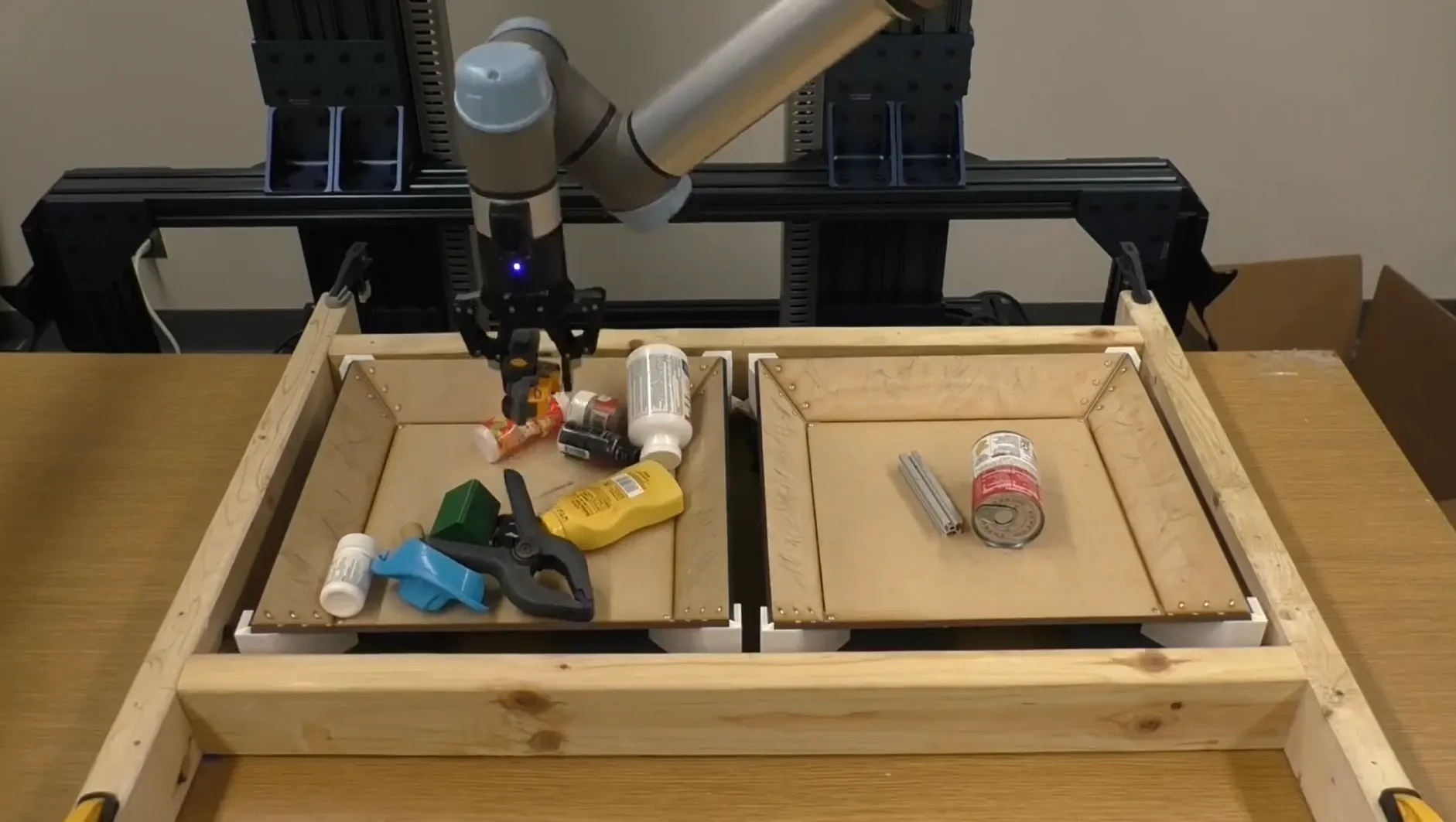 A robot arm picking household objects from a tray, with a yellow bottle labelled as the grasp target.